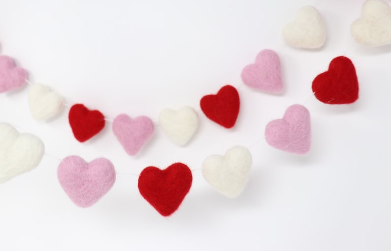 May include: A garland of felt hearts in pink, red, and white. The hearts are strung together with white thread and are hanging against a white background.