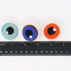 Peut inclure: Trois d&eacute;corations oculaires en laine feutr&eacute;e ronde de couleur bleu clair, bleu avec un iris marron et orange, chacune avec une pupille noire et un reflet blanc. Une r&egrave;gle noire est plac&eacute;e en dessous, montrant que chaque &oelig;il mesure 2,5 cm.