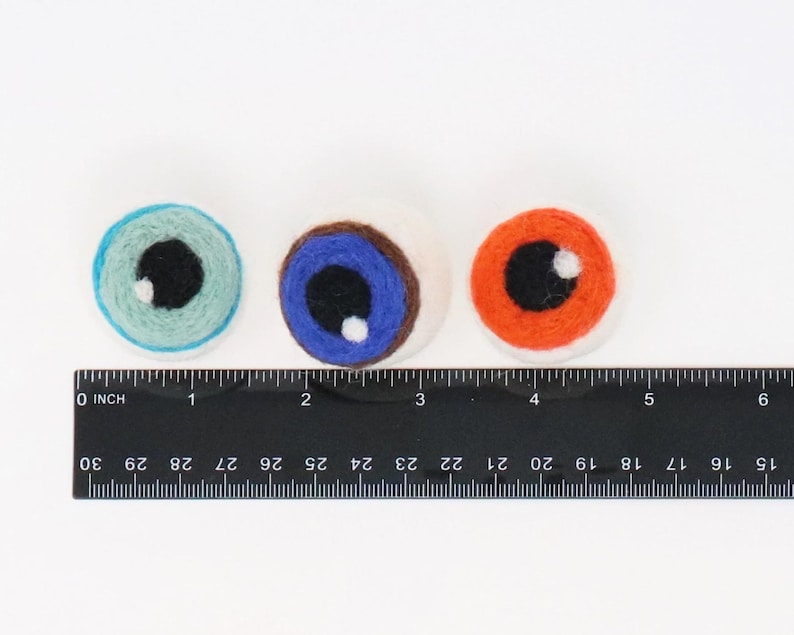 Peut inclure: Trois d&eacute;corations oculaires en laine feutr&eacute;e ronde de couleur bleu clair, bleu avec un iris marron et orange, chacune avec une pupille noire et un reflet blanc. Une r&egrave;gle noire est plac&eacute;e en dessous, montrant que chaque &oelig;il mesure 2,5 cm.