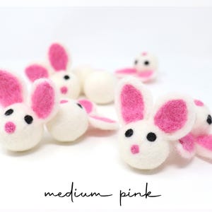 May include: A collection of small, round, white felted wool bunny heads with bright pink inner ears and noses, and black bead eyes. The text medium pink is printed in black script at the bottom of the image against a plain white background.