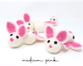 May include: A collection of small, round, white felted wool bunny heads with bright pink inner ears and noses, and black bead eyes. The text medium pink is printed in black script at the bottom of the image against a plain white background.