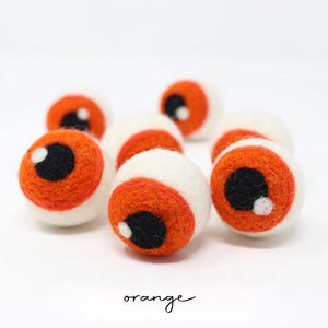 Peut inclure: Un ensemble de sept boules en laine feutr&eacute;e sph&eacute;riques, chacune pr&eacute;sentant une base blanche, un iris orange vif, une pupille noire et un petit point blanc brillant. Le mot orange est imprim&eacute; en texte manuscrit noir en bas de l'image.