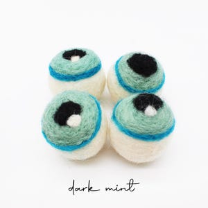 Peut inclure: Quatre boules en laine feutr&eacute;e de forme sph&eacute;rique dispos&eacute;es en carr&eacute;. Chaque boule pr&eacute;sente une base cr&egrave;me, une partie sup&eacute;rieure vert menthe, une bande horizontale bleu vif et un centre noir avec un petit point blanc. Texte : dark mint.