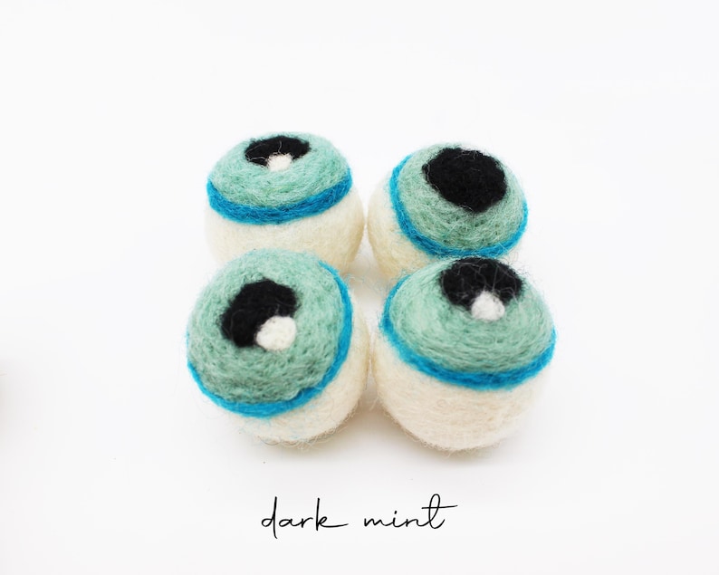 Peut inclure: Quatre boules en laine feutr&eacute;e de forme sph&eacute;rique dispos&eacute;es en carr&eacute;. Chaque boule pr&eacute;sente une base cr&egrave;me, une partie sup&eacute;rieure vert menthe, une bande horizontale bleu vif et un centre noir avec un petit point blanc. Texte : dark mint.