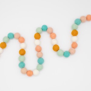Puede incluir: Una guirnalda de bolas de fieltro en tonos melocot&oacute;n, verde menta, blanco y amarillo mostaza. Las bolas est&aacute;n enhebradas en un fondo blanco.