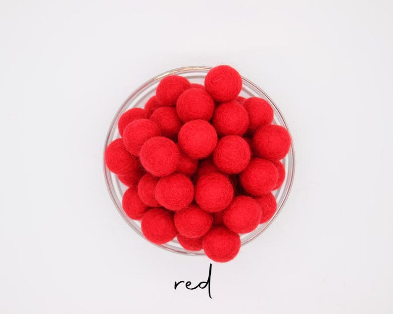 May include: A glass bowl containing many bright red felt balls. The word "red" is written in cursive script beneath the bowl. The balls are uniform in size and closely arranged.