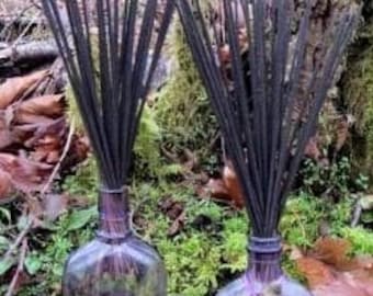 Op de afbeelding: Twee glazen flessen met zwarte wierookstokjes die in de bovenkant zijn gestoken. De flessen zijn helder en hebben een getextureerd oppervlak. De flessen staan op een bed van groen mos.