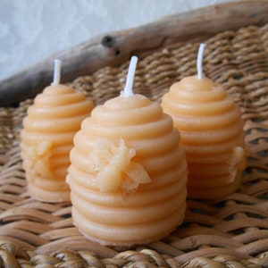 K&ouml;nnte beinhalten: Drei kleine, orangefarbene Bienenwachskerzen in Form von Honigwaben mit einem Bienendesign auf der Vorderseite. Die Kerzen stehen auf einer geflochtenen Weidenoberfl&auml;che.