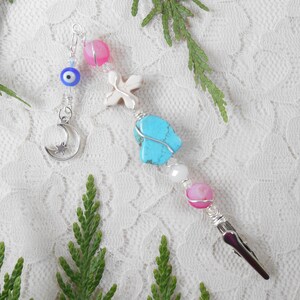 May include: A handmade beaded charm with a silver-tone alligator clip. The charm features a turquoise stone, pink and white beads, a white cross, a blue evil eye, and a silver crescent moon with a star.