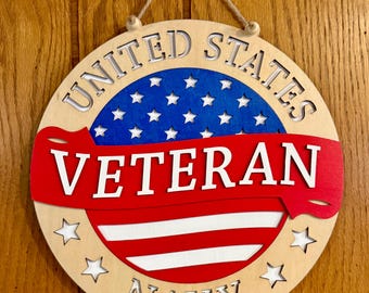 May include: A circular wooden wall sign featuring a blue section with white stars at the top and red and white stripes at the bottom. A red banner across the center reads VETERAN in white letters. The words UNITED STATES and NAVY are cut out.
