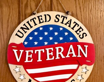 May include: A circular wooden wall sign featuring a patriotic design with a blue section containing white stars, a red banner with the white text VETERAN, and red and white stripes. The text UNITED STATES and MARINES is printed in black.