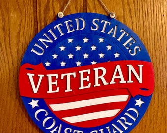 May include: A circular wooden wall sign featuring a blue background with white stars, a red horizontal banner across the center with the white text VETERAN, and the white text UNITED STATES COAST GUARD curved along the top and bottom edges.