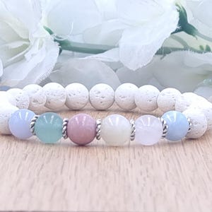 May include: A beaded bracelet featuring white lava rock beads and a series of pastel-colored gemstone beads. The bracelet includes light blue, green, pink, cream, and light pink beads, separated by silver-colored spacers. The bracelet is displayed on a wooden surface.