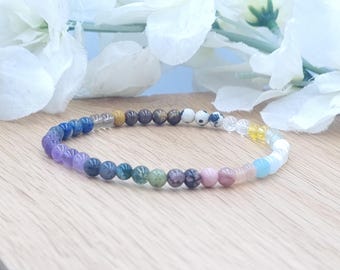 May include: A multi-coloured gemstone bracelet. The bracelet is made up of a variety of beads in shades of purple, blue, green, pink, and yellow. The beads are strung on a clear elastic cord. The bracelet is displayed on a light wood surface.