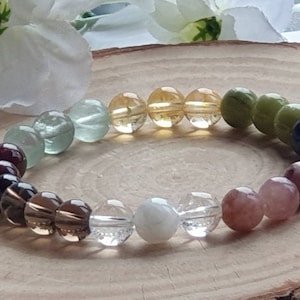 Op de afbeelding: Een kralenarmband met een spectrum aan kleuren, waaronder dieprood, bruin, helder, wit, roze, blauw, groen en geel. De ronde kralen zijn aan elkaar geregen, waardoor een cirkelvormig ontwerp ontstaat. De armband wordt getoond op een houten oppervlak.