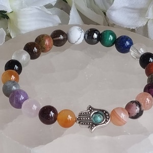 May include: A bracelet made of various colored gemstone beads, including clear, white, brown, red, orange, green, blue, purple, and gray. The bracelet has a silver charm in the shape of a Hamsa hand with a turquoise stone in the center.