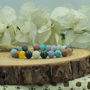 May include: A colourful beaded bracelet with various gemstones. The bracelet includes beads in shades of blue, green, yellow, pink, and white. The bracelet is displayed on a wooden surface with white flowers in the background.