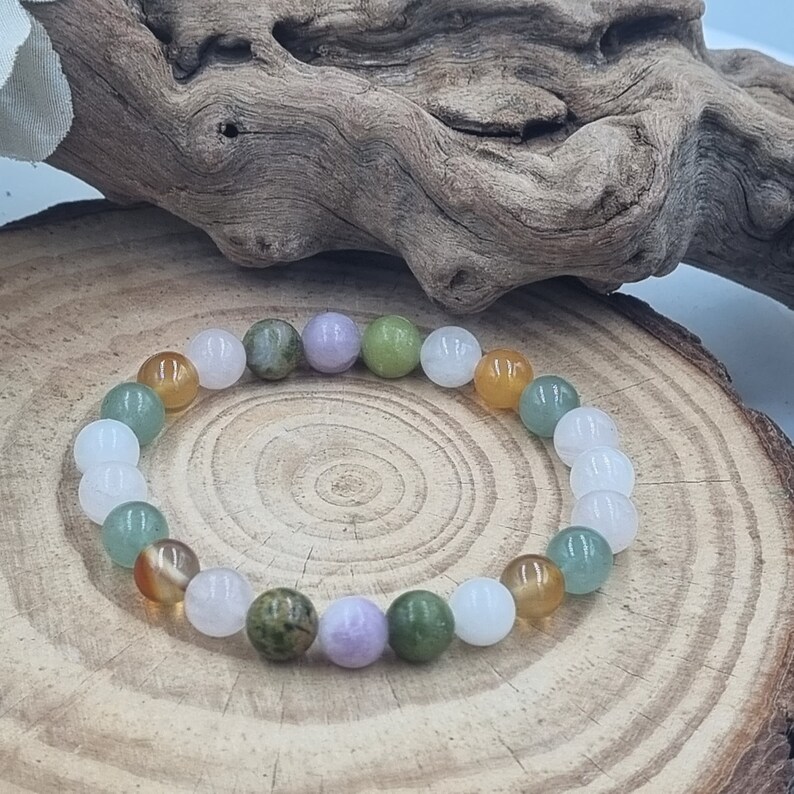 Puede incluir: Un brazalete hecho de cuentas de varios colores, incluyendo verde, blanco, rosa y marr&oacute;n. Las cuentas est&aacute;n enhebradas en un cord&oacute;n el&aacute;stico transparente y est&aacute;n dispuestas en un patr&oacute;n repetitivo. El brazalete est&aacute; sobre una superficie de madera.