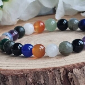 May include: A bracelet made of various colored gemstone beads, including green, blue, purple, orange, white, and black. The bracelet is on a wooden surface.
