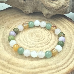 Puede incluir: Un brazalete hecho de cuentas redondas de piedras preciosas de colores. Las cuentas son en tonos de verde, blanco, naranja y morado. El brazalete est&aacute; sobre una superficie de madera.