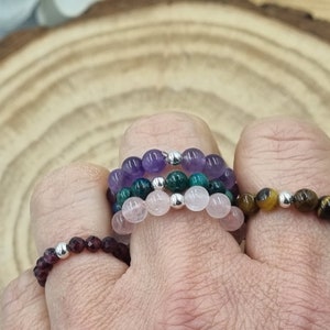 Può includere: Un set di cinque anelli di pietre preziose, ognuno con un colore e una pietra diversi. Gli anelli sono impilati su un dito, con l'anello di ametista in cima, seguito dagli anelli di aventurina verde, quarzo rosa, occhio di tigre e granato. Gli anelli sono realizzati con perline d'argento.