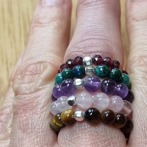 May include: A stack of seven beaded bracelets, each made with different coloured gemstones. The bracelets are stacked on a finger, starting with a dark red garnet bracelet, followed by a green aventurine bracelet, then a purple amethyst bracelet, a pink rose quartz bracelet, and a brown tiger's eye bracelet.
