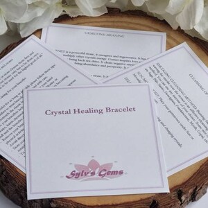 Puede incluir: Una tarjeta blanca con el texto "Crystal Healing Bracelet" y el logotipo "Sylv's Gems" en morado. La tarjeta est&aacute; sobre una superficie de madera con flores blancas en el fondo. La tarjeta tambi&eacute;n incluye informaci&oacute;n sobre la limpieza de piedras preciosas y el significado del granate.