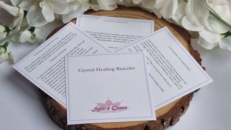 Puede incluir: Una tarjeta blanca con el texto "Crystal Healing Bracelet" y el logotipo "Sylv's Gems" en morado. La tarjeta est&aacute; sobre una superficie de madera con flores blancas en el fondo. La tarjeta tambi&eacute;n incluye informaci&oacute;n sobre la limpieza de piedras preciosas y el significado del granate.