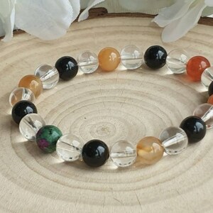 May include: A bracelet made with clear, black, orange, and green gemstone beads. The bracelet is on a wooden surface.
