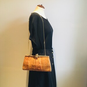 Könnte beinhalten: Eine braune Schlangenleder-Clutch mit einer goldenen Kettenkette und einem goldenen Verschluss. Die Clutch wird von einer Schaufensterpuppe getragen, die ein schwarzes Oberteil und einen schwarzen Rock trägt.