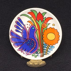Könnte beinhalten: Ein kleiner, weißer Keramikteller mit einem farbenfrohen Design eines blauen Vogels mit rosa und violetten Flügeln, umgeben von grünen, gelben, orangefarbenen und roten Blumen. Der Teller steht auf einem goldenen Metallständer.
