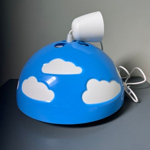 May include: A dome-shaped pendant lamp featuring a bright blue glossy finish decorated with white cloud cutouts. The fixture includes a white ceiling canopy and a white electrical cord extending from the top center of the blue dome structure.
