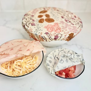 May include: Three bowls covered with reusable food wraps. The wraps are made of fabric and have floral patterns. The bowls contain different foods, including shredded cheese, tomatoes, and an unidentified food.