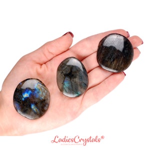 Puede incluir: Tres piedras de labradorita pulidas con superficies lisas y redondeadas. Las piedras muestran tonos gris oscuro y negro con destellos iridiscentes de luz azul y dorada que se reflejan desde la estructura cristalina de los minerales.