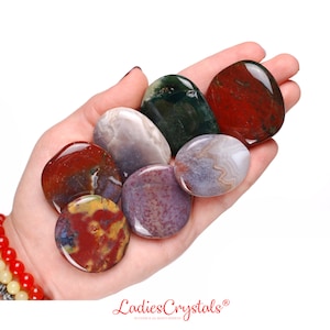 Può includere: Una collezione di sette pietre di agata lisce e levigate, multicolori. Le pietre sono di varie tonalità di rosso, marrone, verde e grigio. Le pietre sono disposte in una mano, con la pietra più grande in alto e la più piccola in basso.