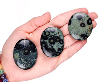 May include: Three green and black oval-shaped polished stones with a unique pattern of black and white spots. The stones are in the palm of a hand.
