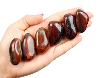 May include: Six smooth, polished red and black banded tumbled stones in a hand. The stones are approximately 1-2 inches in length.