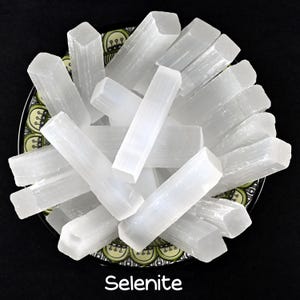 Può includere: Una ciotola piena di bastoncini di selenite bianca. I bastoncini sono lisci e hanno una texture naturale e cristallina. La selenite è un tipo di gesso noto per la sua energia calmante.