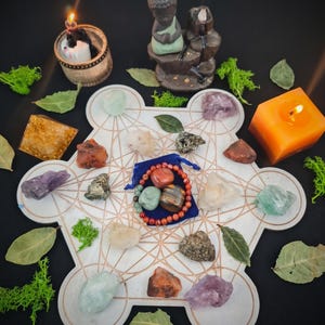 May include: A white geometric grid board featuring copper-colored lines, surrounded by various raw crystals, a blue velvet pouch containing a beaded bracelet, a small Buddha figurine, a pyramid, and lit candles with green moss and dried leaves.
