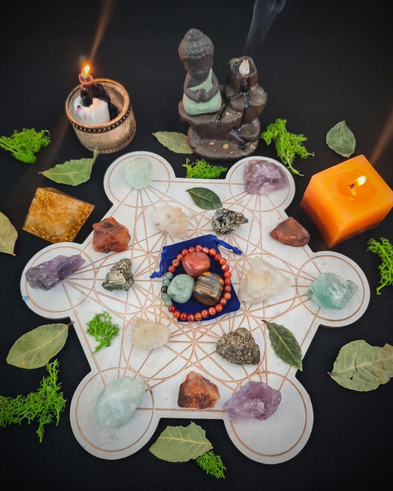 May include: A white geometric grid board featuring copper-colored lines, surrounded by various raw crystals, a blue velvet pouch containing a beaded bracelet, a small Buddha figurine, a pyramid, and lit candles with green moss and dried leaves.