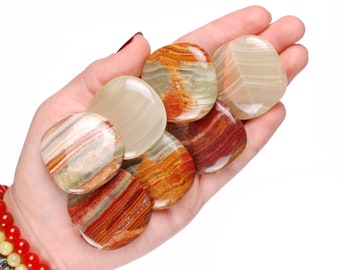 May include: A collection of polished, oval-shaped onyx stones in shades of cream, green, orange, and red. The stones are held in a hand, showcasing their smooth surfaces and natural patterns. The image includes the Ladies Crystals logo.