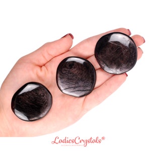 May include: Three polished, round, black obsidian stones with swirling patterns are displayed in an open hand. The stones have a glossy, reflective surface. The image is set against a white background.