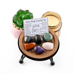 May include: A Kids Sleep Crystal Set with seven polished stones in various colors, displayed on a wooden stand with black metal legs. A small succulent plant in a pink pot and a candle in a glass jar are also present. A card lists the crystals and their properties.