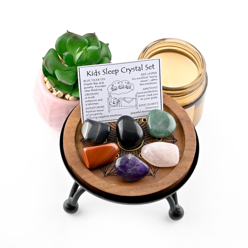 May include: A Kids Sleep Crystal Set with seven polished stones in various colors, displayed on a wooden stand with black metal legs. A small succulent plant in a pink pot and a candle in a glass jar are also present. A card lists the crystals and their properties.