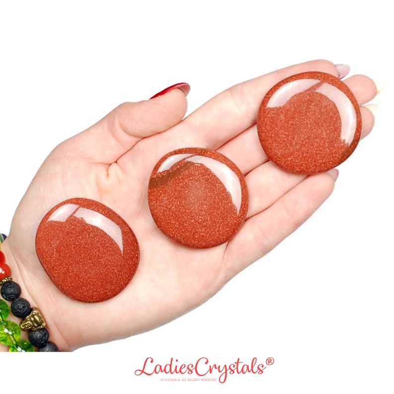 May include: Three polished, round, reddish-brown gemstones with a sparkling, granular texture are displayed in an open hand. The stones have a glossy, reflective surface. The image is set against a white background.