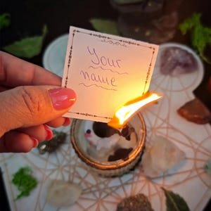 May include: A small white rectangular paper with the handwritten text Your name in purple ink, held over a burning candle flame. The paper features a thin black decorative border with intricate corner details and is being consumed by fire.