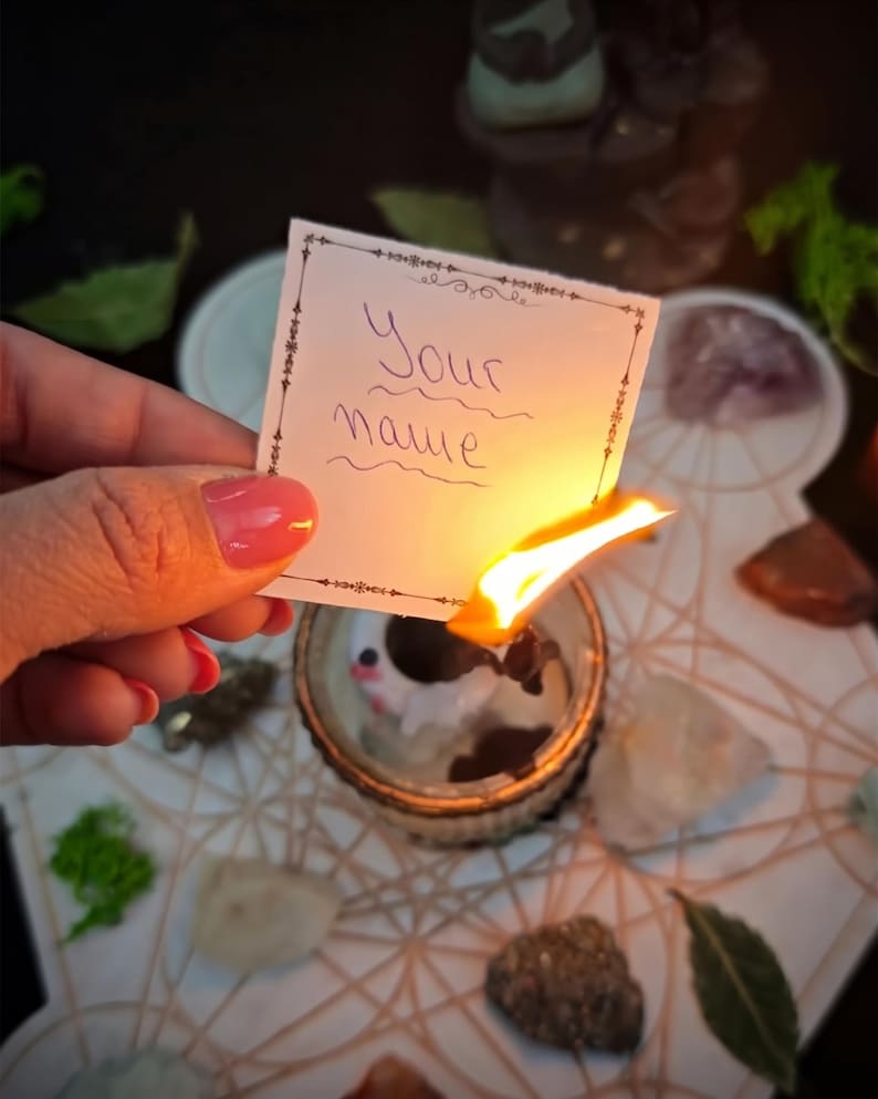 May include: A small white rectangular paper with the handwritten text Your name in purple ink, held over a burning candle flame. The paper features a thin black decorative border with intricate corner details and is being consumed by fire.