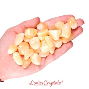 May include: A hand holding a pile of yellow-orange tumbled citrine stones. The stones are smooth and polished.