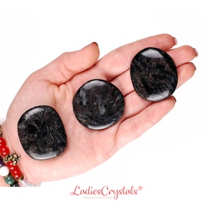 May include: Three smooth, round, black and grey gemstone palm stones. The stones are in the palm of a hand.