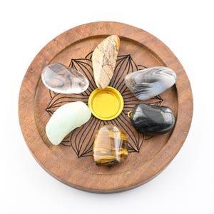 May include: A wooden tray with a floral design holds seven polished gemstones of various colors and patterns. The gemstones include clear, brown, green, black, and tiger's eye stones. A gold-colored circular object is in the center.